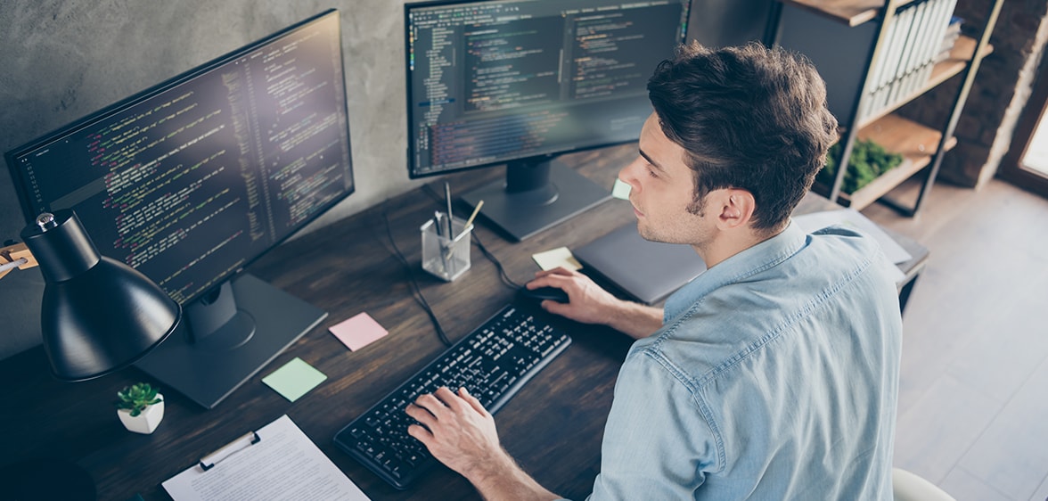 Egy programozó ül egy íróasztalnál és két monitor van előtte amin dolgozik.